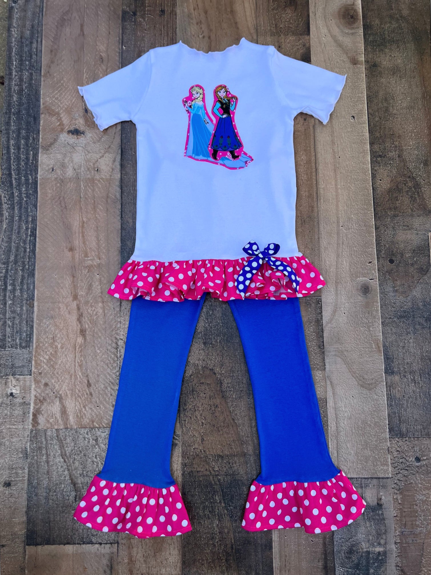 A children's two-piece outfit laid flat on a rustic wood plank background. The set includes a light blue short-sleeve top with a colorful Frozen graphic on the chest and a pink polka-dot ruffle hem, paired with bright blue flared leggings that have matching pink polka-dot ruffle hems and a blue-and-white polka-dot bow at the waist.