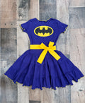 A children's short-sleeve dress in royal blue with a yellow Batman logo on the chest, a yellow waist bow, and yellow trim along the ruffled hem, laid flat on a rustic wood plank background.