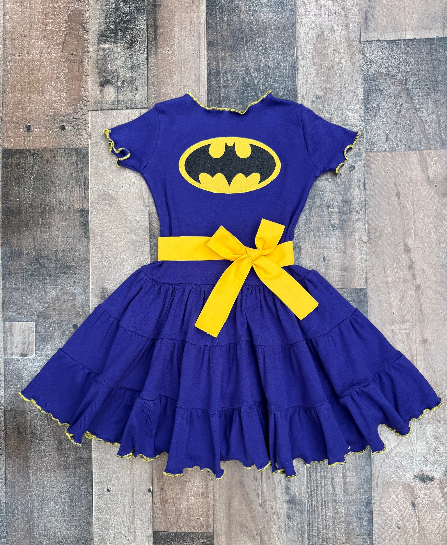 A children's short-sleeve dress in royal blue with a yellow Batman logo on the chest, a yellow waist bow, and yellow trim along the ruffled hem, laid flat on a rustic wood plank background.